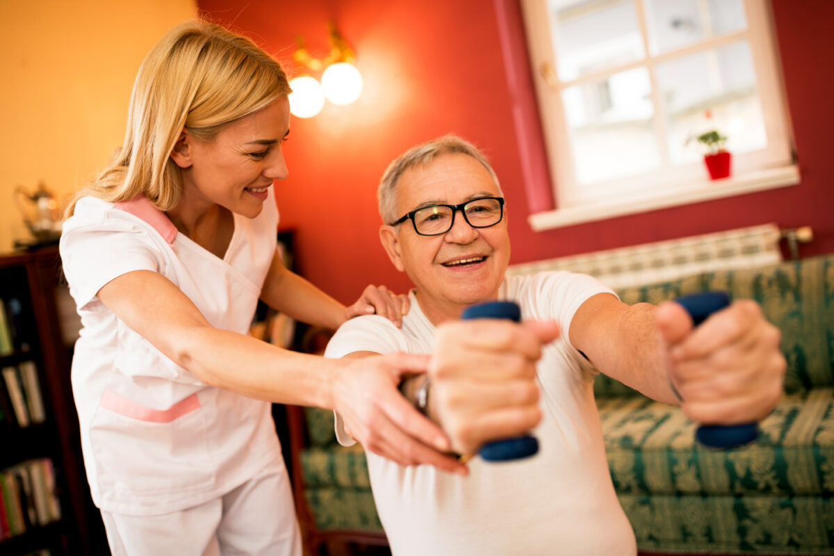 Senior mature man doing exercises at an Assisted Living with dumbbells while nurse helps to them