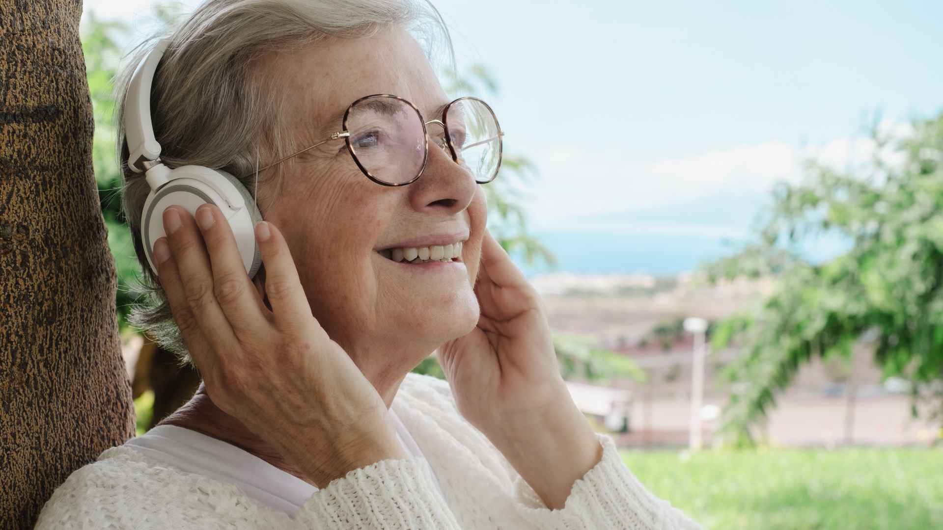Elderly smiling woman enjoys a peaceful moment outdoors sitting against a tree listening music