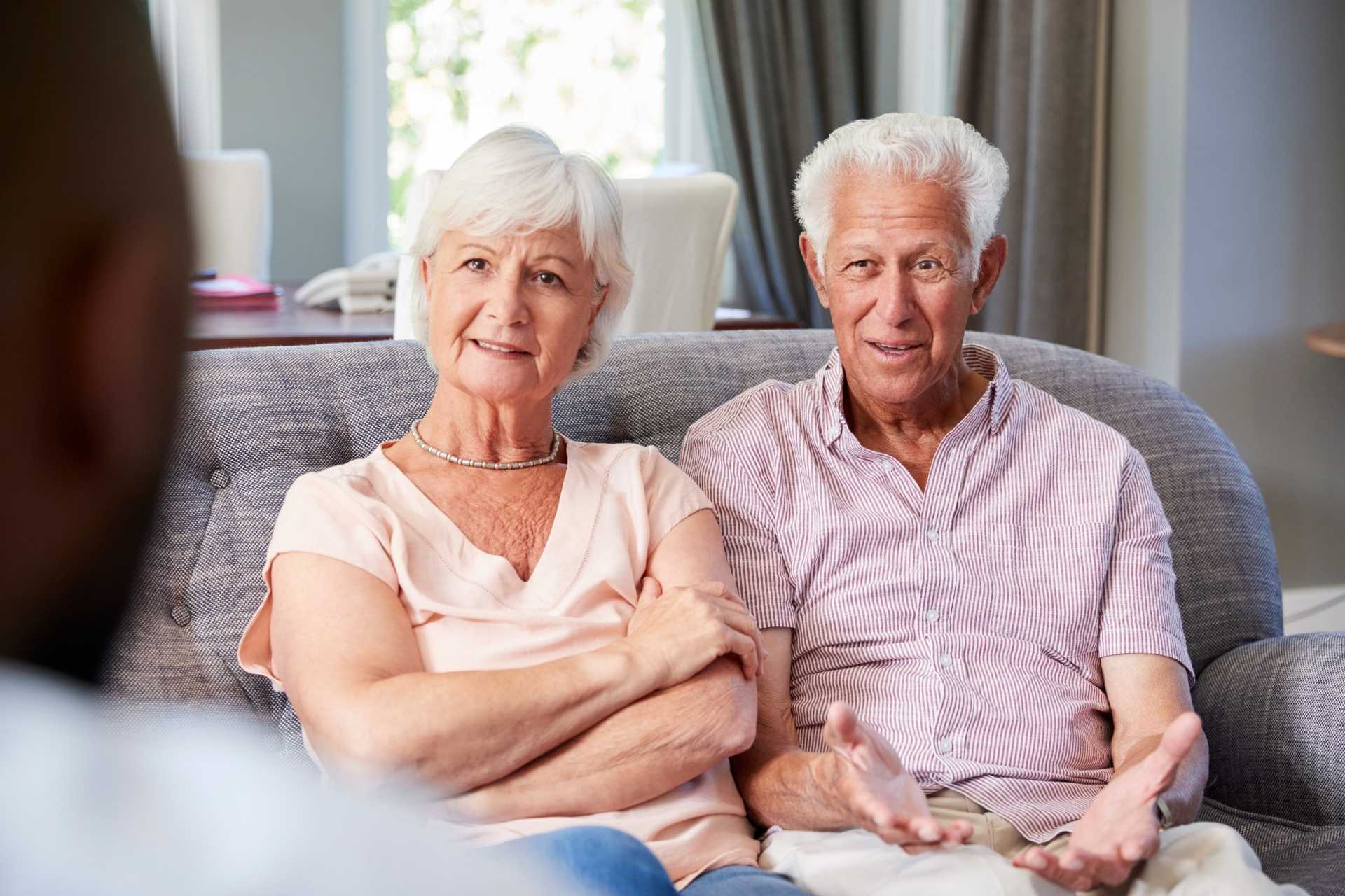 Two seniors sitting together on a couch listening to someone.